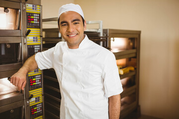 Male baker in chef uniform leaning against oven rack smiling in bakery kitchen with ovens, trays