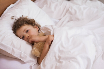 Girl with curly hair wearing pajamas hugging teddy bear lying on bed in bedroom, copy space
