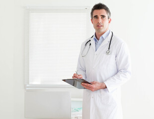 Male doctor standing in exam room holding clipboard and writing notes with stethoscope, copy space