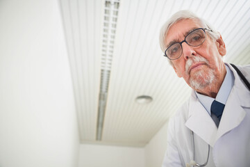 Senior doctor leaning toward viewer in exam room wearing lab coat with stethoscope, copy space