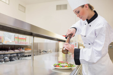 Female chef wearing white coat seasoning salad with pepper mill at kitchen counter, copy space