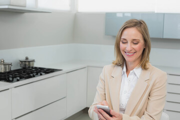 Businesswoman smiling while holding smartphone checking messages near stove in kitchen, copy space