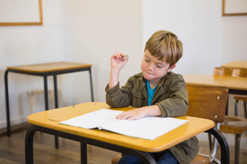 School-age boy sitting at desk in classroom, focusing on workbook holding pencil wearing jacket