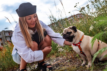 Cute little tan and black pug breed of dog on a walk with harness and a black leash walking with in the city park. Street photography. The owner is walking his pet.	