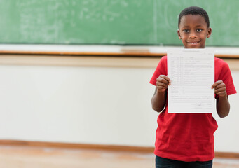 African American boy holding completed worksheet in classroom before green chalkboard, copy space