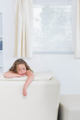 Female child leaning over back of white leather sofa in bright living room with window blinds