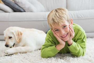 Blond boy lying on white shag rug at home smiling while puppy chewing bone near sofa