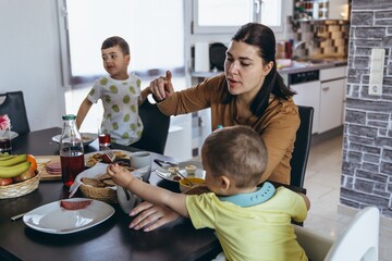 Mother having breakfast with two young sons at home kitchen. Toddler in high chair reaching for...