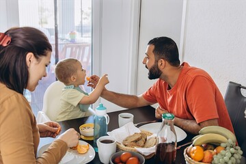 Happy parents having breakfast with toddler son at home. Father gently feeding baby at the table....