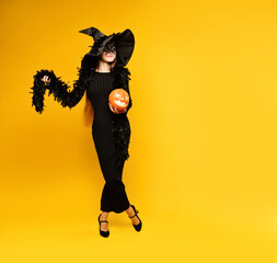 Cheerful young woman witch wearing black dress, hat, carnival mask holds pumpkin jack o lantern on yellow background. Concept halloween. Girl in witch costume.
