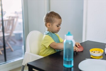 Happy baby boy in one year sitting in a high chair wearing a silicone bib. He is eating while a blue baby water bottle stands on the table in the foreground