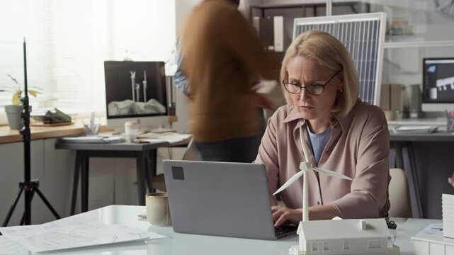 Medium time lapse of busy middle-aged blonde Caucasian female engineer working on laptop at green energy company, while male colleagues are discussing solar battery specifications in background