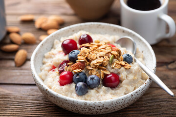 Oatmeal porridge bowl with berries, crunchy granola topping, cup of black coffee on rustic wooden table background, closeup view. Healthy breakfast meal