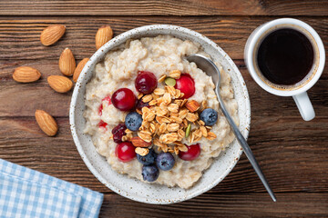 Oatmeal porridge bowl with berries, granola topping and cup of black espresso coffee on a wooden table background, top view