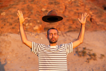 Man in striped shirt with raised hands and floating hat at golden hour outdoors