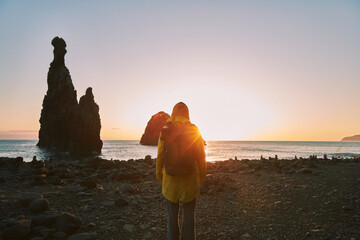 Traveler in yellow jacket stands on a rocky beach, gazing at majestic sea stacks silhouetted...