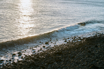 Sunlight glistening on gentle ocean waves as they meet a dark, pebbled beach, creating a serene and...