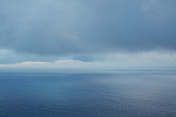 Vast serene blue ocean meets a dramatic, moody sky filled with heavy grey clouds, creating an expansive, tranquil natural seascape scene stretching to the distant horizon.