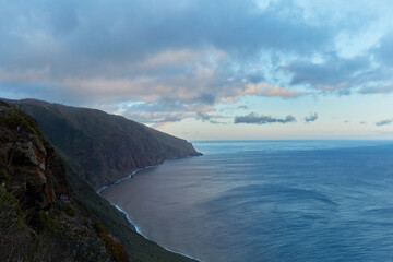 Panoramic view of a rugged coastline meeting the vast ocean under a dramatic cloudy sky at dusk, showcasing the natural beauty of a steep cliffside landscape.