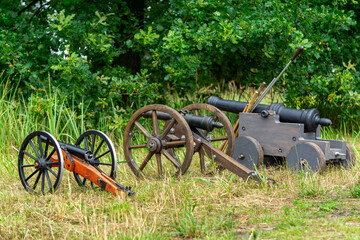 Replicas of old cannons with wooden wheels placed on grass, set against a green background. Scene...