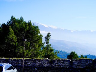 Scenic mountain view with snow-capped peaks, pine trees, and a stone wall in the foreground under a...