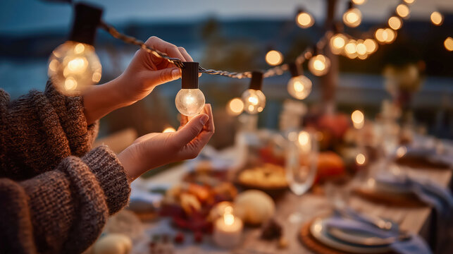 Woman's hands adjusting glowing string light bulbs for a cozy outdoor Thanksgiving day dinner setting, soft warm light