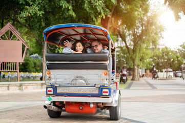 Old white man and asian woman waving a hand while riding in tuk tuk three wheel taxi touring a city.