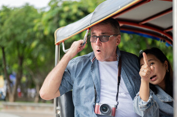 Old white man and asian woman looking around while riding in tuk tuk three wheeled taxi touring city