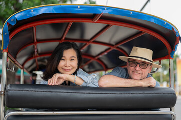Old white man and asian woman looking back while riding in a tuk tuk three wheel taxi touring a city