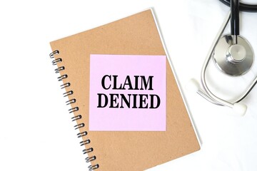 Claim Denied on a sticky note above a notebook. Bright white background and doctor's stethoscope.