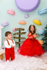 A little girl and a boy in red clothes have fun in the artificial snow by the fireplace in the studio. The girl is holding a box with gifts. Little kids in red christmas costume having fun beside a