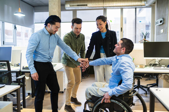 Multi ethnic businesspeople joining hands in modern coworking office, showing support for disabled colleague in wheelchair, promoting inclusion and teamwork