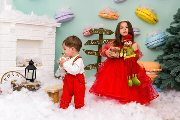 A little girl and a boy in red clothes have fun in the artificial snow by the fireplace in the studio. The girl is holding a box with gifts. Little kids in red christmas costume having fun beside a