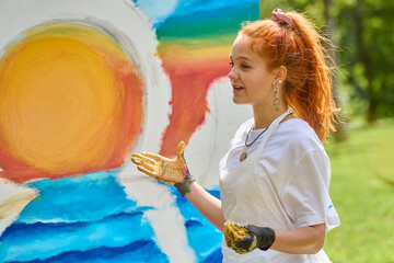 Young female with red hair discusses vibrant ocean sunset painting outdoors. Wearing white apron,...