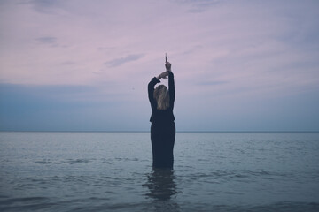 Woman stands waist-deep in tranquil ocean at sunset, raising her arms in contemplation. Peaceful...