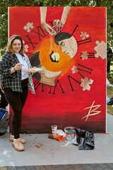 Young adult female with light brown hair outdoors, standing beside large red surreal painting with puzzle motifs. Dominant theme is time and abstraction