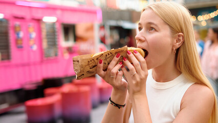 Young woman with long blonde hair enjoying hot dog topped with cheese and toppings at vibrant...