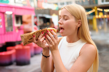 Young woman with long blonde hair enjoying a tasty hot dog wrapped in decorated paper at colorful...