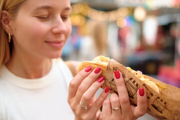 Close-up of woman with closed eyes and gentle smile holding large hot dog wrapped in vintage print...