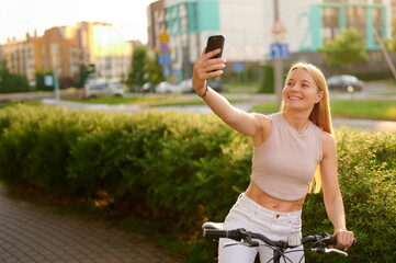 woman takes selfie with smartphone while cycling through urban park during golden hour. casual...