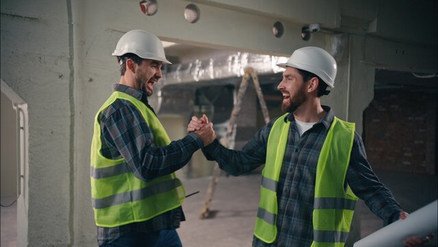 Two happy Caucasian men greeting friends buddies males guys workers in uniform helmets business construction teamwork engineers architects meeting shaking hands building cooperation colleagues gesture
