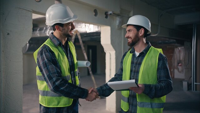 Two glad Caucasian men builders discuss building project in construction architects handymen talking paper draft workmen guys working document manufactory contractors smile shake hands deal handshake