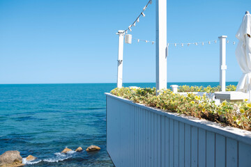 A tranquil seaside restaurant terrace with white wooden furniture and a bed of yellow flowers,...