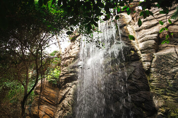 A stunning waterfall cascades down rugged rocks, surrounded by lush green foliage. Sunlight filters...