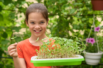 White girl in her 10s smiling and holding tray of mung bean sprouts in green garden. Concept of sustainable gardening, healthy food, and childhood education