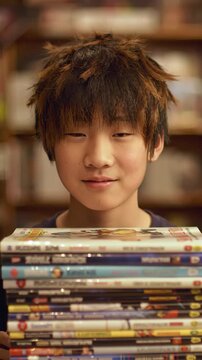  Happy Young Asian Boy with Messy Hair Proudly Holding a Large Stack of DVDs or Manga in a Library