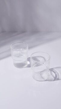 Glass cups and a jug of clean, clear drinking water stand on a white tabletop. Sunny day and sharp shadows from glass