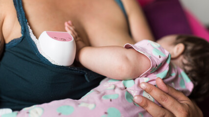 Close-up of mother breastfeeding baby while using electric breast pump for milk expression,...