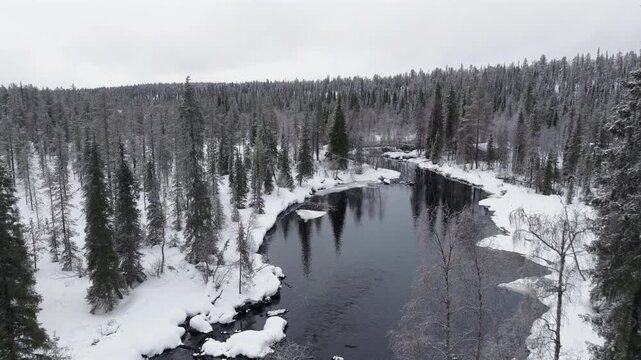 Aerial, flying backwards above a river surrounded by snow-covered forest