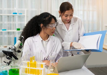 Two female scientists in lab coats are collaborating in a laboratory. One scientist with curly hair and safety goggles using laptop and microscope while other standing and pointing to science document